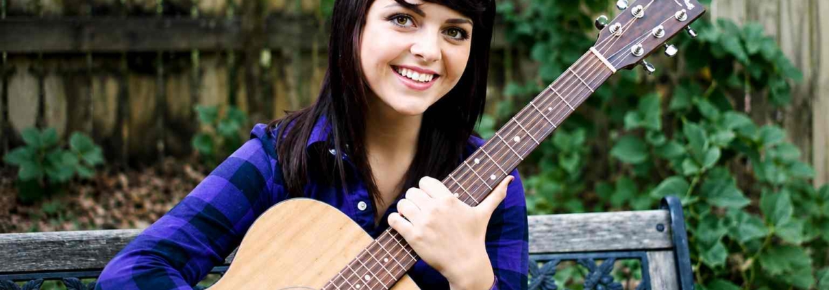 Young woman with dark hair smiling while holding an acoustic guitar outdoors on a bench with a wooden fence and green plants in the background.