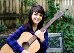 Young woman with dark hair smiling while holding an acoustic guitar outdoors on a bench with a wooden fence and green plants in the background.