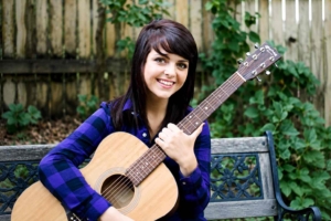 Young woman with dark hair smiling while holding an acoustic guitar outdoors on a bench with a wooden fence and green plants in the background.