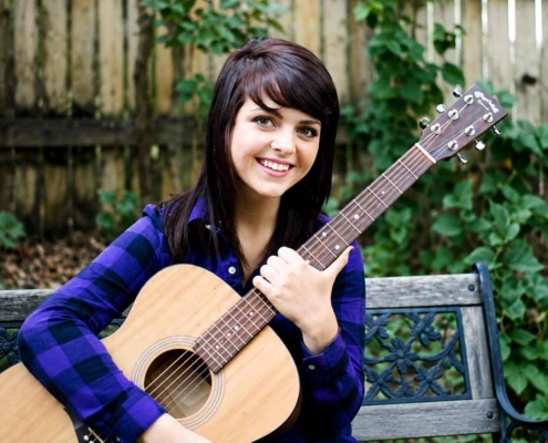 Young woman with dark hair smiling while holding an acoustic guitar outdoors on a bench with a wooden fence and green plants in the background.