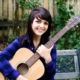 Young woman with dark hair smiling while holding an acoustic guitar outdoors on a bench with a wooden fence and green plants in the background.