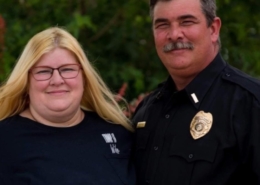Harry Bailey standing outdoors beside his wife while wearing a black uniform shirt with a badge and clerical collar.