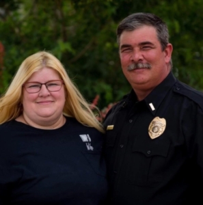 Harry Bailey standing outdoors beside his wife while wearing a black uniform shirt with a badge and clerical collar.