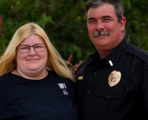 Harry Bailey standing outdoors beside his wife while wearing a black uniform shirt with a badge and clerical collar.