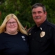 Harry Bailey standing outdoors beside his wife while wearing a black uniform shirt with a badge and clerical collar.