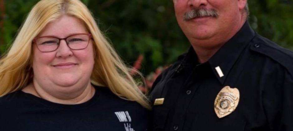 Harry Bailey standing outdoors beside his wife while wearing a black uniform shirt with a badge and clerical collar.