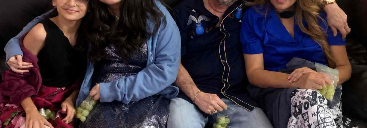 A smiling family of four sitting closely together on a dark leather couch indoors, dressed in semi-formal clothing and holding green grapes in a cozy home setting.