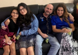 A smiling family of four sitting closely together on a dark leather couch indoors, dressed in semi-formal clothing and holding green grapes in a cozy home setting.