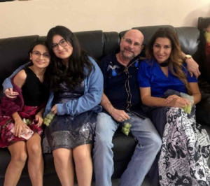 A smiling family of four sitting closely together on a dark leather couch indoors, dressed in semi-formal clothing and holding green grapes in a cozy home setting.