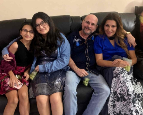 A smiling family of four sitting closely together on a dark leather couch indoors, dressed in semi-formal clothing and holding green grapes in a cozy home setting.