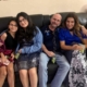 A smiling family of four sitting closely together on a dark leather couch indoors, dressed in semi-formal clothing and holding green grapes in a cozy home setting.
