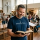 Male Christian minister wearing a navy “MINISTER” t-shirt and silver cross necklace while reading an open Bible inside a modern church gathering space with people in the background.