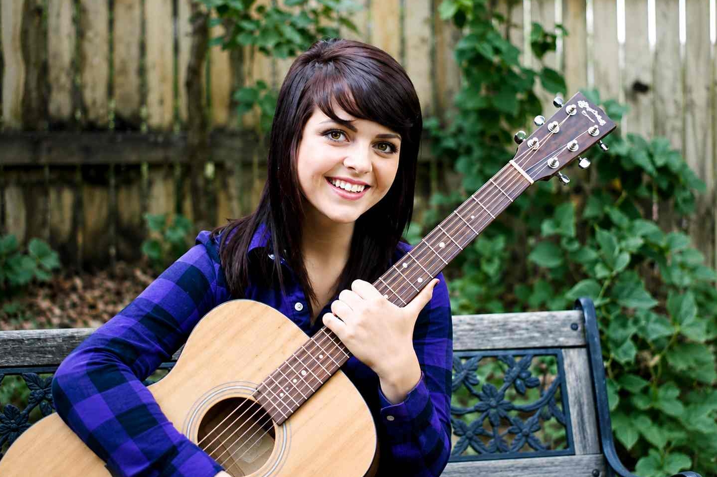 Young woman with dark hair smiling while holding an acoustic guitar outdoors on a bench with a wooden fence and green plants in the background.